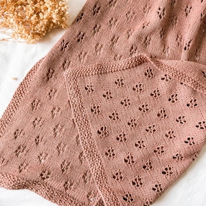 May include: A dusty rose knitted blanket featuring a repeating openwork diamond pattern and a textured garter stitch border. The fabric displays a consistent knit structure with small, evenly spaced holes throughout the textile surface.