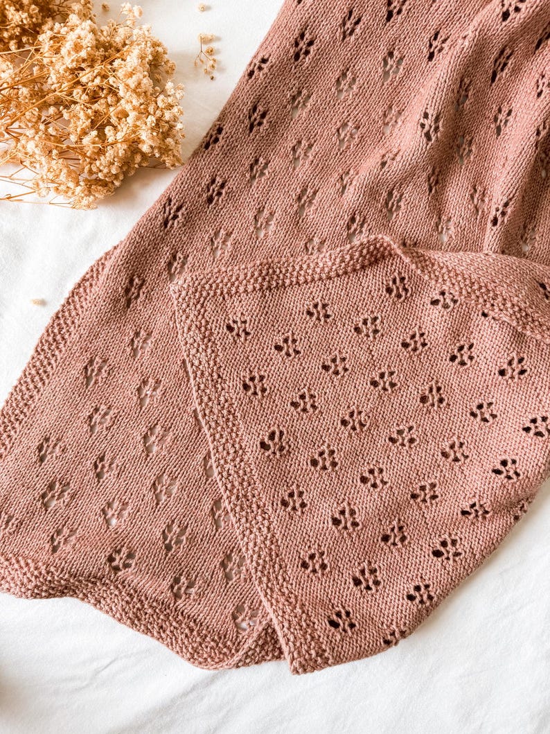 May include: A dusty rose knitted blanket featuring a repeating openwork diamond pattern and a textured garter stitch border. The fabric displays a consistent knit structure with small, evenly spaced holes throughout the textile surface.