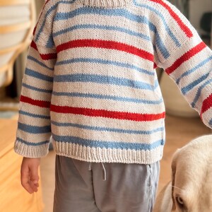 Pode incluir: Um su&eacute;ter de malha com listras horizontais em azul claro, vermelho e branco sujo. O su&eacute;ter tem gola redonda e mangas compridas. As mangas e o corpo s&atilde;o listrados. O su&eacute;ter &eacute; usado com cal&ccedil;as cinzentas.