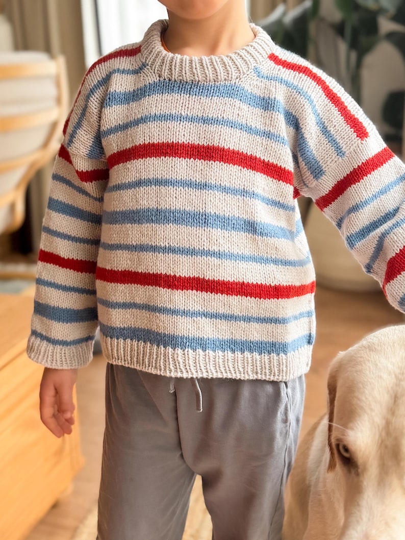 Pode incluir: Um su&eacute;ter de malha com listras horizontais em azul claro, vermelho e branco sujo. O su&eacute;ter tem gola redonda e mangas compridas. As mangas e o corpo s&atilde;o listrados. O su&eacute;ter &eacute; usado com cal&ccedil;as cinzentas.
