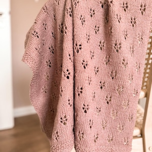 May include: A dusty rose knitted blanket featuring a repeating eyelet lace pattern throughout the fabric. The textile has a textured surface with a consistent knit stitch and a simple border along the edges, showcasing a soft, muted pink colour.