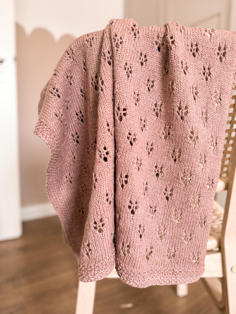 May include: A dusty rose knitted blanket featuring a repeating eyelet lace pattern throughout the fabric. The textile has a textured surface with a consistent knit stitch and a simple border along the edges, showcasing a soft, muted pink colour.