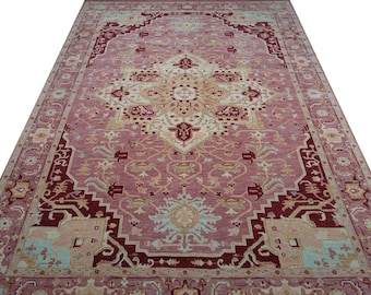 May include: A large, rectangular rug with an intricate, symmetrical design. The rug features a central medallion in cream and gold, surrounded by a field of muted rose. The border is a deep burgundy with geometric patterns and accents of light blue and cream.