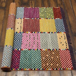 May include: A colorful, rectangular rug with a geometric pattern. The rug features a patchwork design with various colors including blue, pink, yellow, and red. The rug is partially rolled up, revealing the patterned underside.