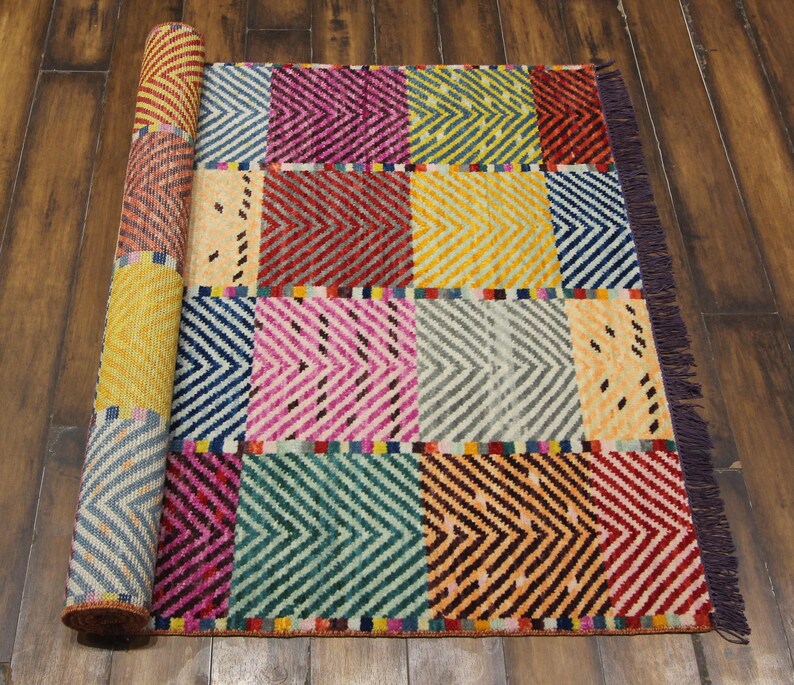 May include: A colorful, rectangular rug with a geometric pattern. The rug features a patchwork design with various colors including blue, pink, yellow, and red. The rug is partially rolled up, revealing the patterned underside.