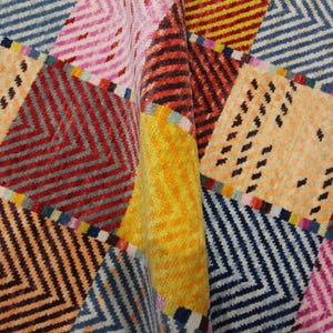 May include: A colorful, patterned textile with a geometric design. The design features a patchwork of squares and rectangles in various colors, including orange, red, blue, pink, and yellow. The pattern includes zigzags and small black dashes.