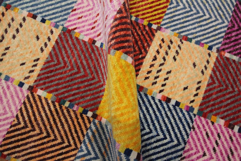 May include: A colorful, patterned textile with a geometric design. The design features a patchwork of squares and rectangles in various colors, including orange, red, blue, pink, and yellow. The pattern includes zigzags and small black dashes.