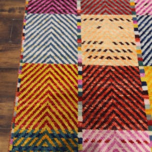 May include: A colorful rug with a geometric pattern. The rug features a patchwork design with squares of various colors, including yellow, blue, red, and pink. Each square has a chevron pattern. The rug is made of a soft, textured material.