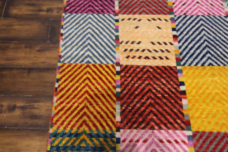 May include: A colorful rug with a geometric pattern. The rug features a patchwork design with squares of various colors, including yellow, blue, red, and pink. Each square has a chevron pattern. The rug is made of a soft, textured material.