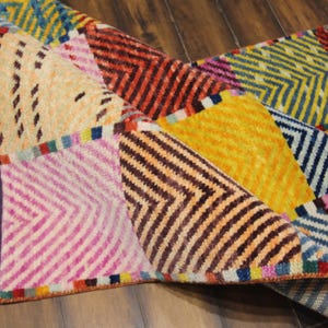 May include: A colorful, patterned rug with a geometric design. The rug features a variety of colors, including pink, yellow, orange, and blue. The rug has a fringed edge and is made of a woven material.