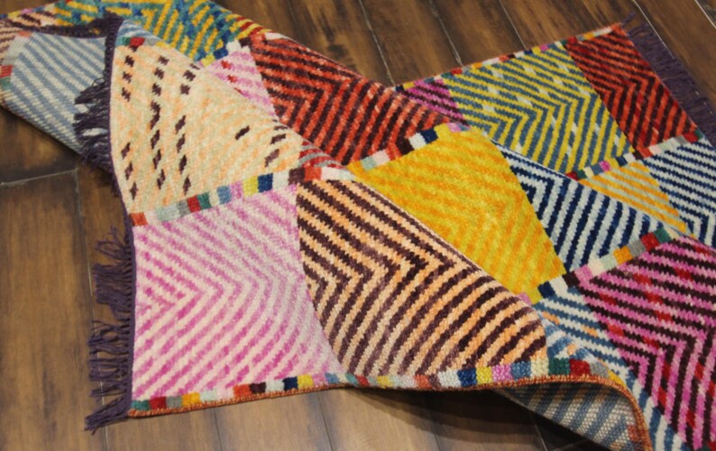 May include: A colorful, patterned rug with a geometric design. The rug features a variety of colors, including pink, yellow, orange, and blue. The rug has a fringed edge and is made of a woven material.