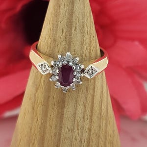 以下が含まれることがあります： 中央に楕円形のルビーの宝石があり、小さな透明なアクセントストーンで囲まれた金の指輪。指輪には、中央の石の両側にダイヤモンド型のディテールが付いた金のバンドがあります。指輪は木の円錐の上に展示されています。