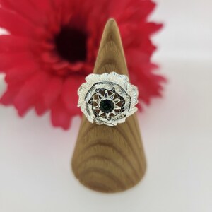 May include: A silver ring with a green gemstone in the center, surrounded by a decorative design of leaves. The ring is displayed on a wooden ring holder.