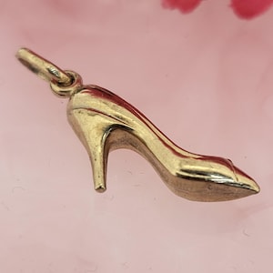 May include: A gold-coloured charm in the shape of a high-heeled shoe. The charm has a pointed toe and a slender heel. The shoe charm is suspended from a small loop, suggesting it can be added to a bracelet or necklace.