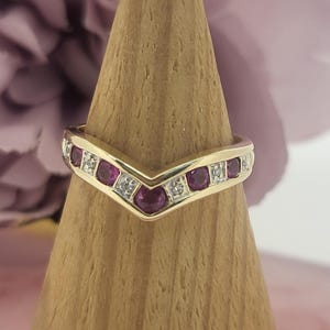 May include: A gold ring with a V-shaped design. The ring features alternating round, deep red gemstones and small, clear gemstones. The ring is displayed on a wooden, cone-shaped stand against a soft pink background.