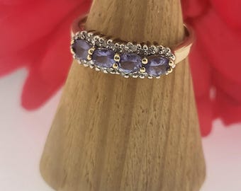May include: A gold ring featuring five oval, light purple gemstones set in a row. The ring is displayed on a wooden cone-shaped stand, with a blurred red background.