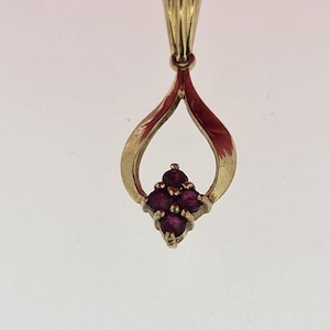 May include: A gold pendant with a teardrop shape. The pendant has a cluster of small, round, deep red gemstones at the base. The top has a textured loop for a chain. A classic jewellery piece.