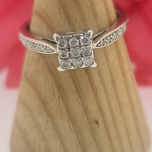 May include: A silver ring featuring a square cluster of small, clear stones. The band is also set with smaller stones. The ring is displayed on a wooden ring holder, with a blurred pink background.