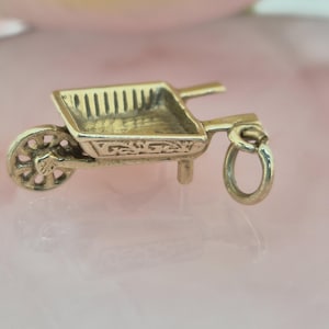 May include: A gold-toned metal charm shaped like a miniature wheelbarrow. The charm features a single spoked wheel at the front, a rectangular basin with a textured interior, two handles at the rear, and a small circular jump ring for attachment.