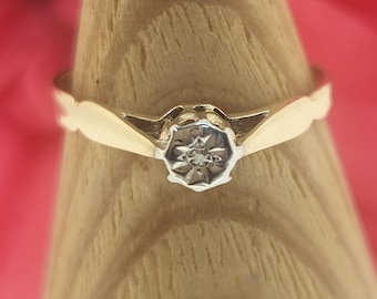 Könnte beinhalten: Ein goldener Ring mit einem Sternmuster und einem kleinen Diamanten in der Mitte. Der Ring wird auf einem hölzernen Ringhalter präsentiert. Das Band hat ein gewelltes Design.