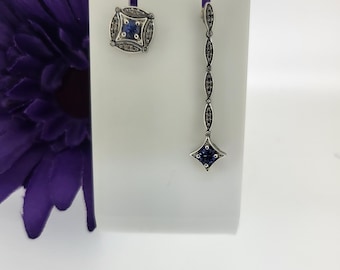 May include: A pair of silver earrings with blue gemstones. One earring is a square with a blue gemstone in the center, surrounded by smaller clear gemstones. The other earring is a long, thin dangle with a blue gemstone at the bottom.