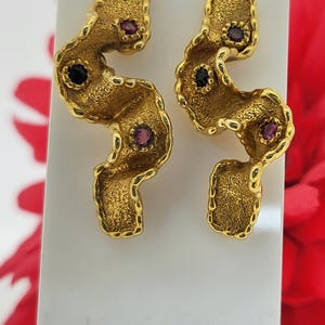 May include: A pair of gold-toned earrings with a textured, organic design. Each earring features a wavy, abstract shape with embedded gemstones: two dark blue and two red. The earrings are displayed on a white surface.
