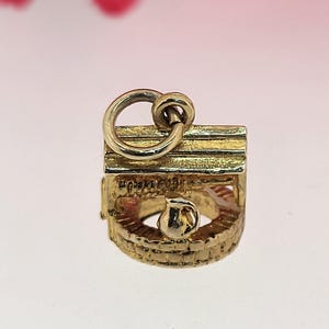 May include: A gold-colored charm in the shape of a well, with a small jug inside. The charm has a detailed brickwork design and a loop at the top for attaching to a bracelet or necklace. The well charm is a classic jewelry piece.