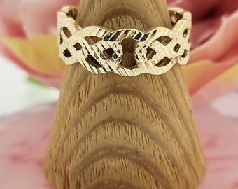 May include: A gold ring featuring a repeating openwork pattern. The ring is presented on a wooden, cone-shaped stand, set against a soft pink and floral backdrop. The ring's detailed design is clearly visible.