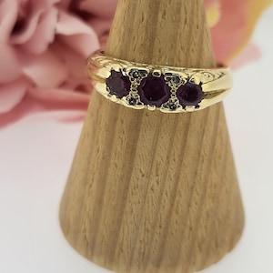 May include: A gold band ring with three round, dark purple gemstones set in the centre. The ring is on a wooden display stand.