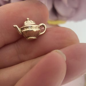 Pode incluir: Um pequeno pingente de chaleira dourada, com tampa, bico e alça. A chaleira em miniatura é muito detalhada e parece ser feita de metal. O pingente é segurado na mão de uma pessoa, com um fundo desfocado.