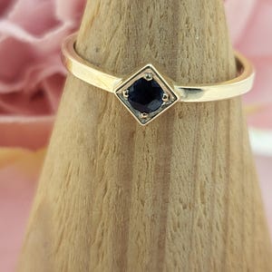 Op de afbeelding: Een gouden ring met een vierkante, donkerblauwe edelsteen gezet in een ruitvormige zetting. De ring wordt gepresenteerd op een houten ringhouder tegen een zachtroze achtergrond. De band is dun en gepolijst.