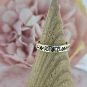 May include: A gold band ring featuring alternating round red rubies and clear white diamonds, each set within a starburst engraving. The ring is displayed on a wooden cone stand, showcasing the polished metal finish and the detailed star-shaped settings.