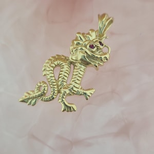 Könnte beinhalten: Ein goldener Drachenanhänger mit strukturiertem, geschupptem Körper und einem detaillierten Kopf mit zwei kleinen, runden, dunkelrosa Edelsteinaugen. Der Anhänger verfügt über eine polierte Goldöse an der Oberseite zur Befestigung.