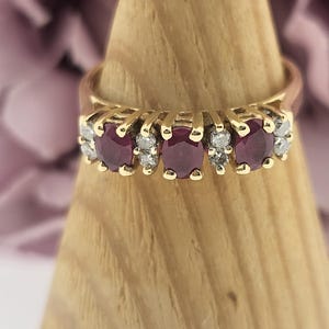 Może przedstawiać: Złoty pierścionek z pięcioma owalnymi rubinami i małymi diamentami. Rubiny są w kolorze głębokiej czerwieni, a diamenty są przezroczyste. Pierścionek jest prezentowany na drewnianym stojaku z rozmytym kwiatowym tłem.