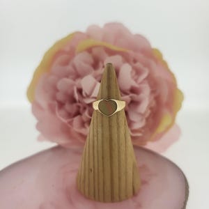 Könnte beinhalten: Ein goldfarbener Ring mit herzförmigem Gesicht. Der Ring wird auf einem kegelförmigen Holzständer präsentiert, vor einem weichen rosa Hintergrund mit einer verschwommenen Blume. Das Herz des Rings hat ein dezentes Streifenmuster.