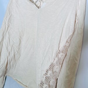 May include: A long-sleeved, cream-colored shirt with a V-neck and lace detailing. The back and one sleeve feature intricate lace patterns, adding a delicate touch to the casual top. The shirt is made of a soft, lightweight fabric.