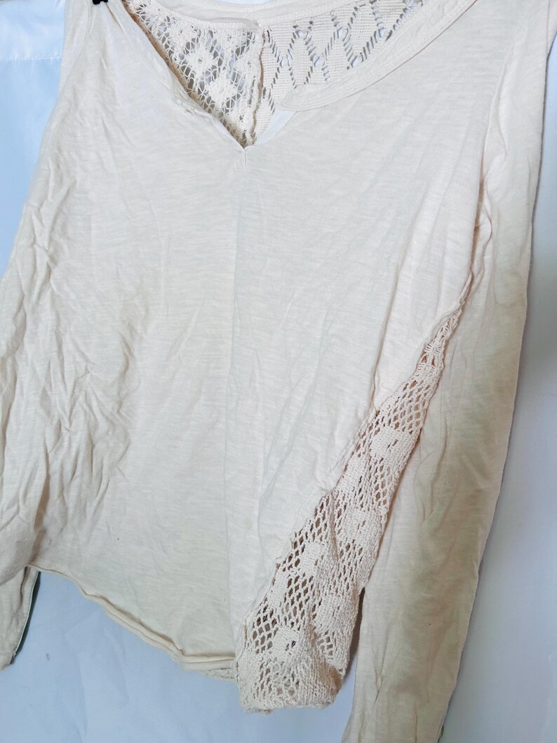 May include: A long-sleeved, cream-colored shirt with a V-neck and lace detailing. The back and one sleeve feature intricate lace patterns, adding a delicate touch to the casual top. The shirt is made of a soft, lightweight fabric.