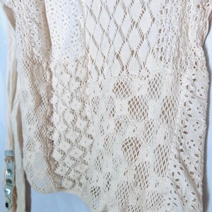 May include: Cream-colored long-sleeved top with intricate lace and openwork details. The design features various patterns, including diamond and floral motifs. The sleeve cuff has decorative buttons.
