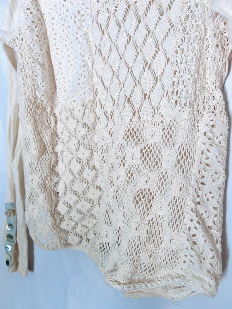 May include: Cream-colored long-sleeved top with intricate lace and openwork details. The design features various patterns, including diamond and floral motifs. The sleeve cuff has decorative buttons.