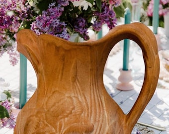 May include: A tan ceramic pitcher featuring a raised floral relief design on the front surface. The pitcher has a wide, curved handle and a flared rim. The body of the vessel displays a textured, wood-grain-like pattern surrounding the flower.