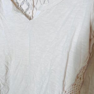 May include: Cream-colored V-neck shirt with lace detailing. The shirt features a delicate lace panel across the upper back and along the side seam, adding a touch of texture and visual interest to the soft, flowing fabric. A casual top.