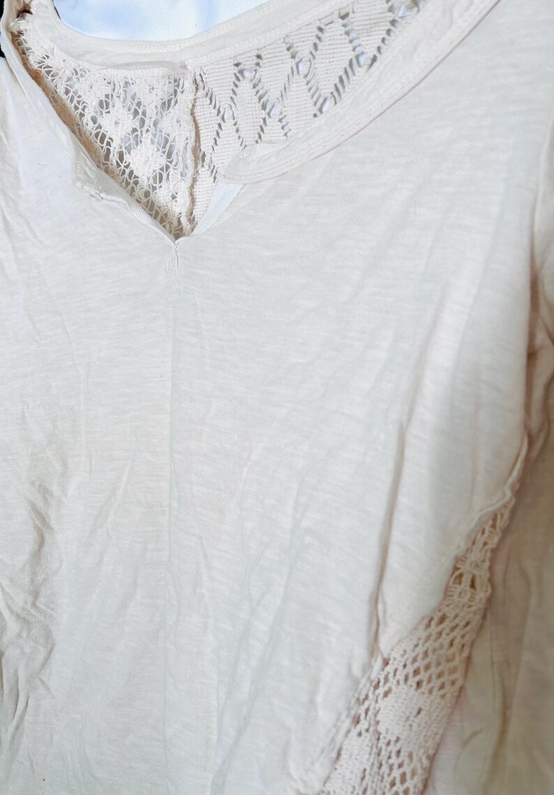 May include: Cream-colored V-neck shirt with lace detailing. The shirt features a delicate lace panel across the upper back and along the side seam, adding a touch of texture and visual interest to the soft, flowing fabric. A casual top.