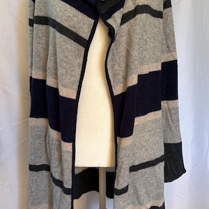 May include: A hooded cardigan in a striped pattern. The open-front sweater features horizontal stripes in shades of gray, navy blue, and a light beige. The sleeves and hood are also striped. The cardigan is displayed on a mannequin.
