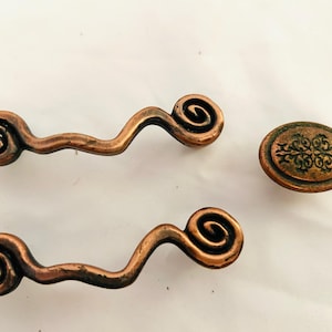 May include: Three antique copper-colored cabinet hardware pieces. Two are wavy pulls with spiral ends, and one is a round knob with an embossed floral design. The hardware has a distressed finish.