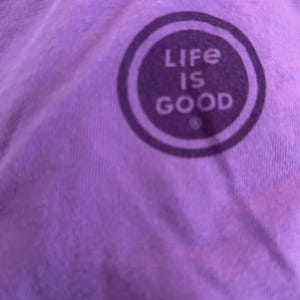 May include: Purple t-shirt with a white circle logo that says "Life is Good" in all caps.