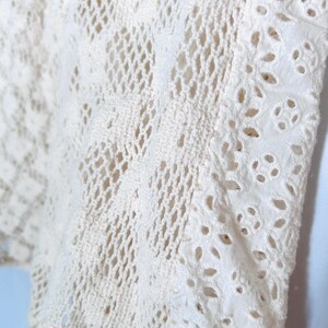 May include: Close-up of a cream-colored, open-weave fabric. The fabric features a combination of crochet and cutwork details. The crochet sections have a diamond pattern, while the cutwork has floral-shaped cutouts. The edges are finished with a decorative trim.