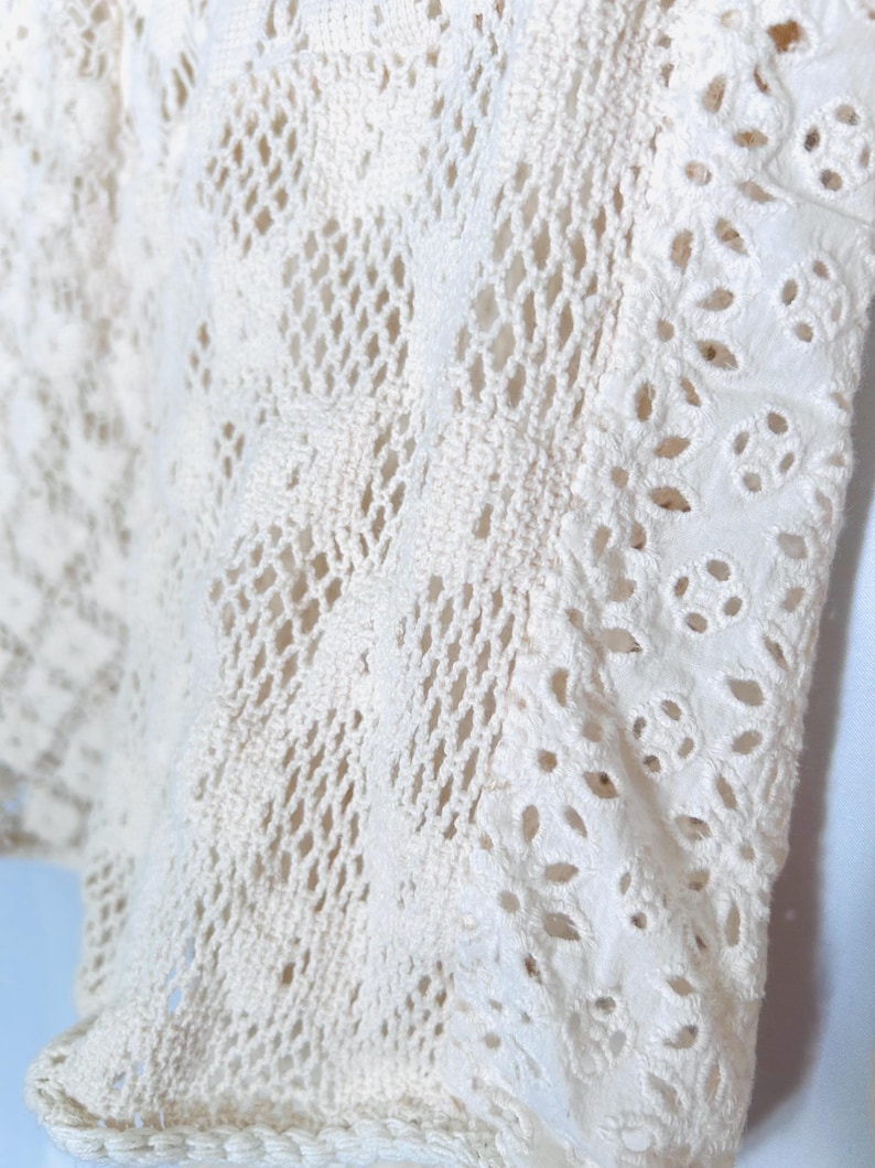 May include: Close-up of a cream-colored, open-weave fabric. The fabric features a combination of crochet and cutwork details. The crochet sections have a diamond pattern, while the cutwork has floral-shaped cutouts. The edges are finished with a decorative trim.