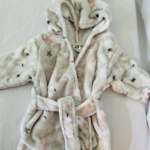 May include: A light gray hooded robe with a matching belt. The robe is decorated with pink and black star and floral patterns. The robe is made of a soft, plush material.