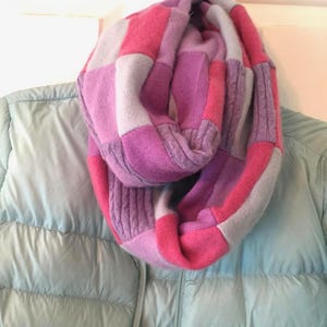 May include: A pink, purple, and light blue knitted infinity scarf. The scarf is made of a soft, chunky yarn and has a checkered pattern.