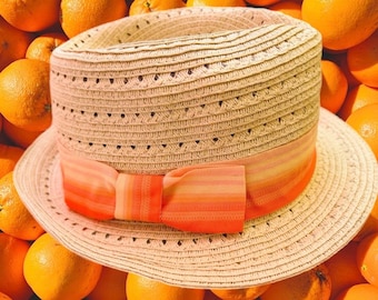 May include: A beige straw fedora hat featuring a woven texture with small triangular ventilation holes. The hat is accented by a wide, horizontal striped ribbon band in shades of orange, coral, and yellow, finished with a decorative bow.